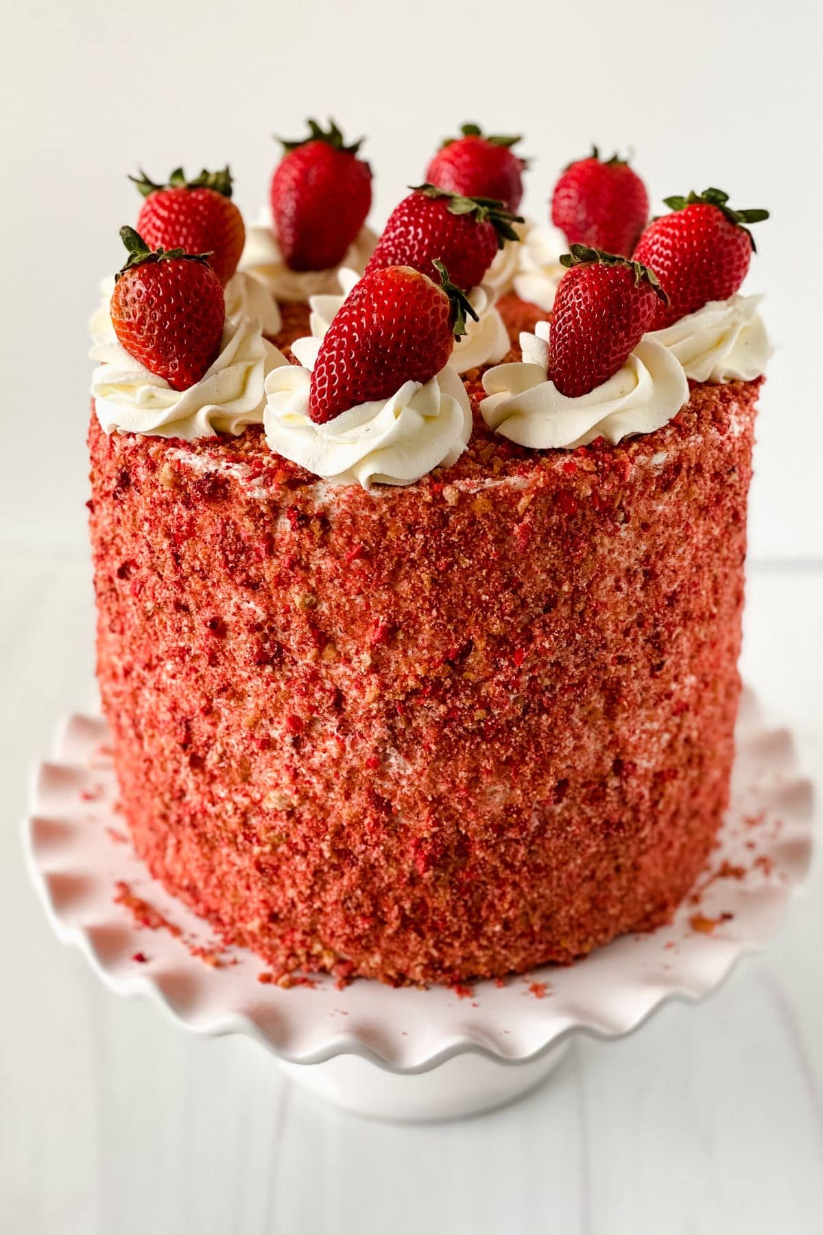 A tall Strawberry Crunch Cake Recipe creation covered in red crumbs, topped with swirls of white frosting and whole strawberries, displayed on a white scalloped cake stand.