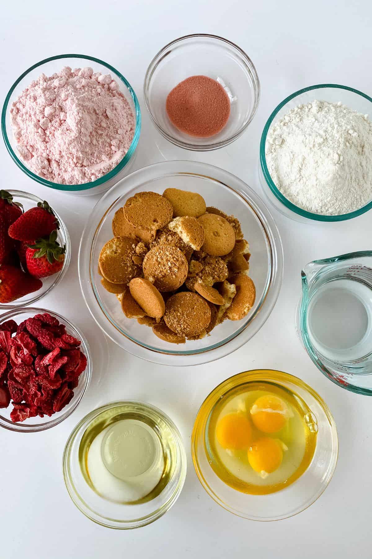 Top-down view of baking ingredients in bowls for a Strawberry Crunch Cake Recipe, including flour, eggs, oil, water, strawberries, freeze-dried fruit, strawberry powder, and vanilla wafer cookies on a white surface.