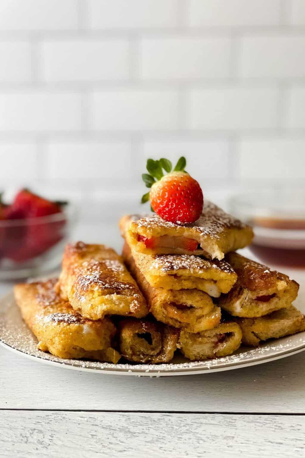 A plate of recipe filled with fruit, dusted with powdered sugar, and topped with a fresh strawberry. A bowl of strawberries and a dipping sauce are in the background.