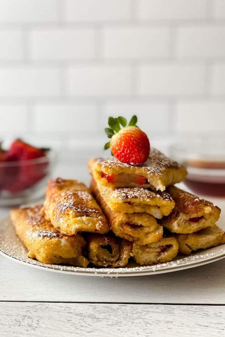 A plate of recipe filled with fruit, dusted with powdered sugar, and topped with a fresh strawberry. A bowl of strawberries and a dipping sauce are in the background.