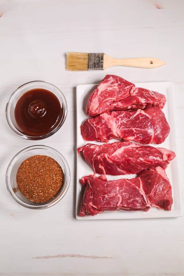 Four ingredients on a rectangular plate next to bowls of sauce and seasoning, with a brush on a white surface.