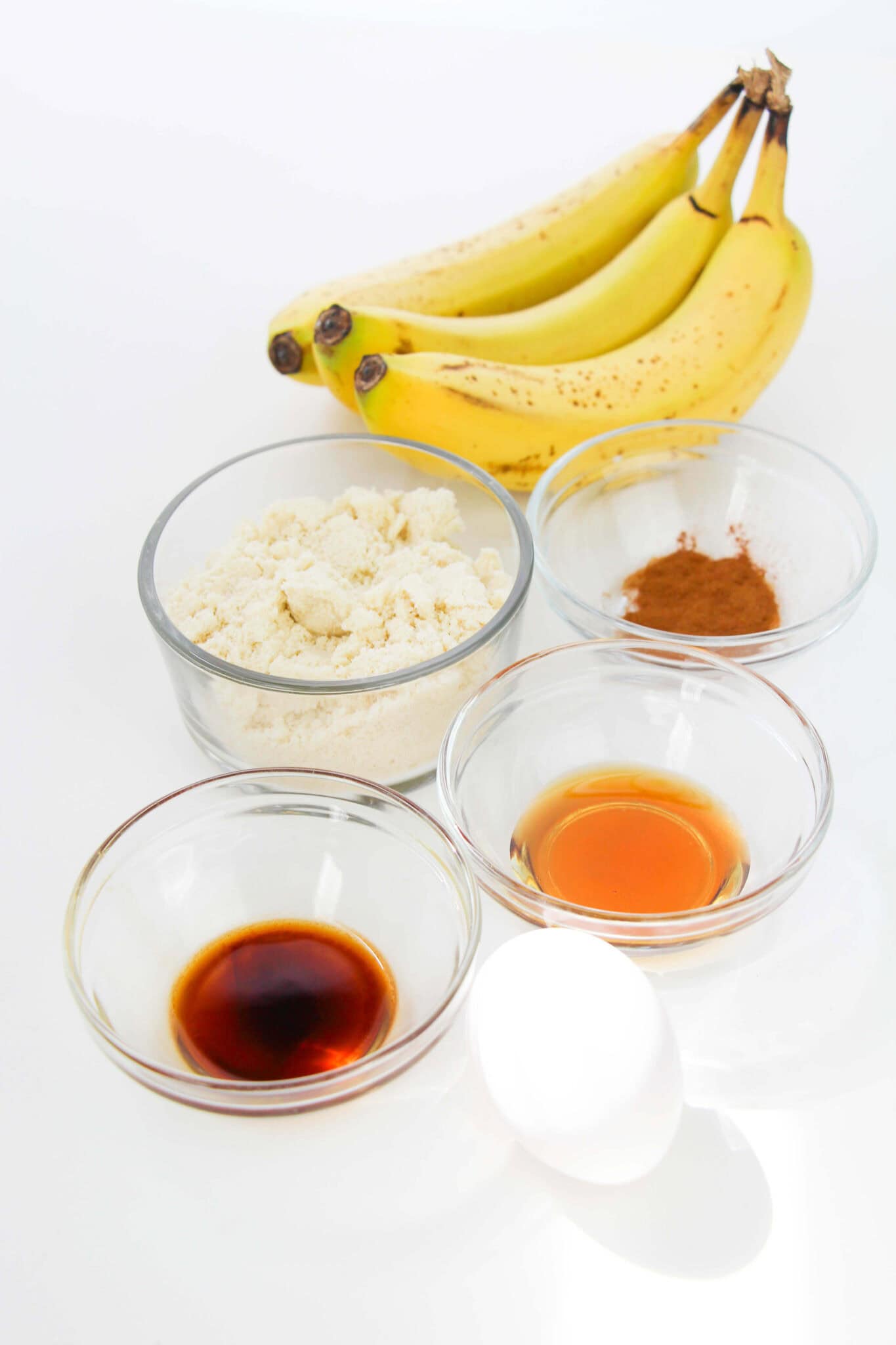 Ingredients for delicious banana bread cookies: ripe bananas, flour, cinnamon, vanilla extract, honey, and an egg arranged on a pristine white surface.