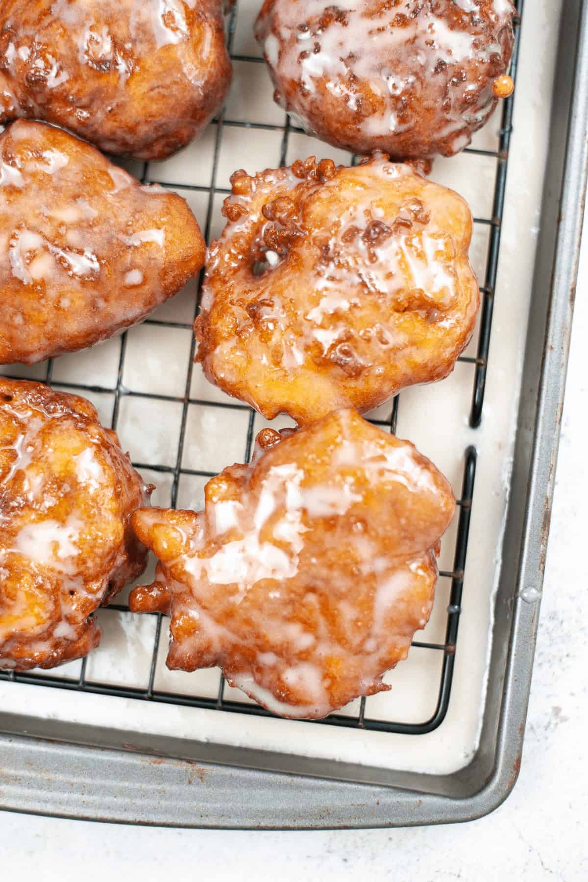 Close-up of homemade apple fritters, perfectly glazed, resting on a cooling rack over a baking sheet.