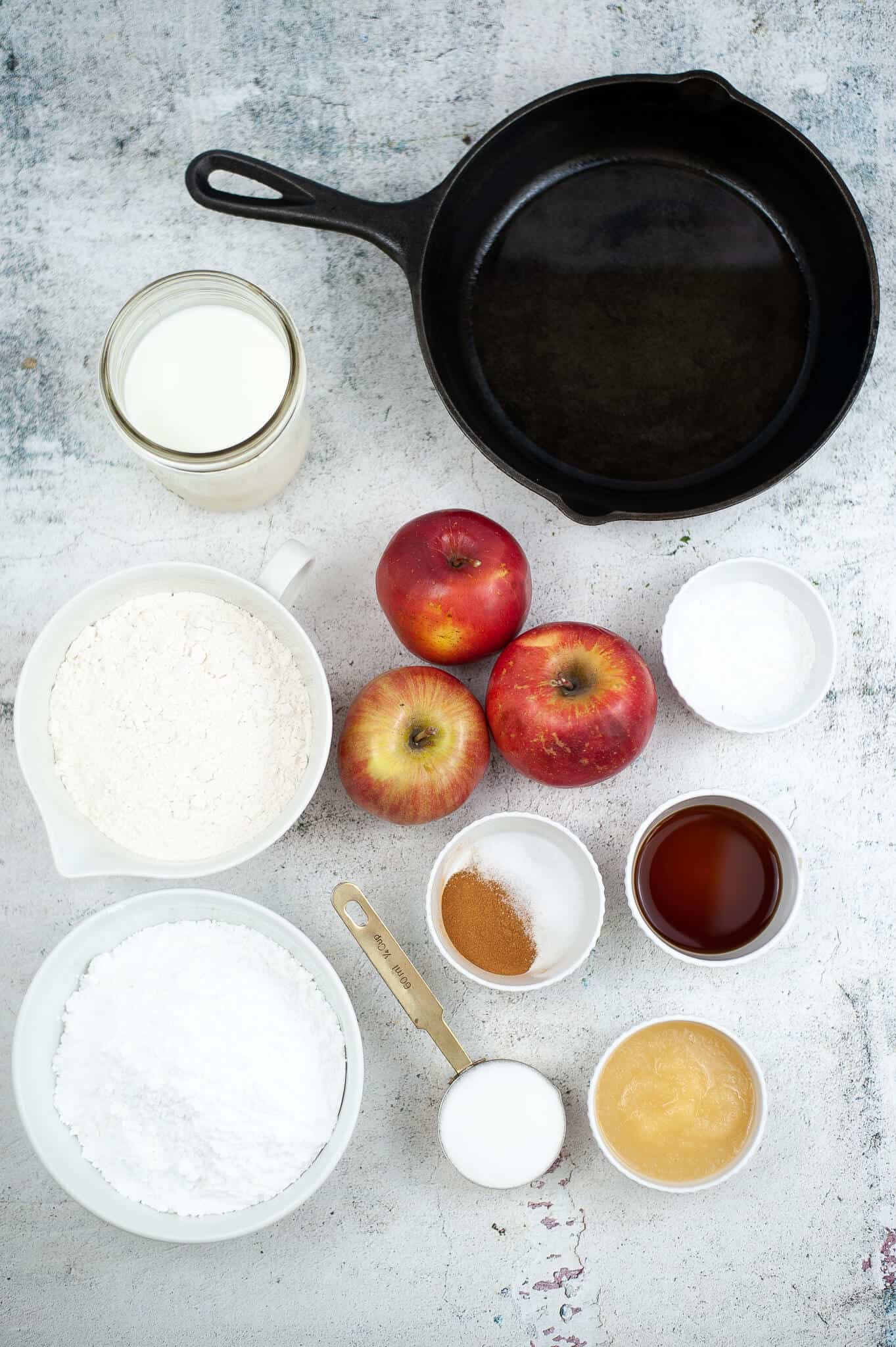 Ingredients laid out for baking homemade apple fritters: a skillet, milk, apples, flour, baking soda, baking powder, cinnamon, vinegar, sugar, applesauce, and powdered sugar on a textured surface.