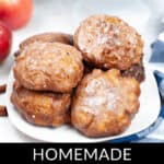A plate of homemade apple fritters glistens with a sweet glaze, while fresh apples sit enticingly in the background.