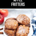 A plate of homemade apple fritters rests invitingly on the table, surrounded by fresh apples, aromatic cinnamon sticks, and a charming checkered napkin.