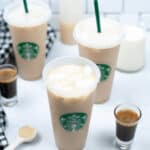 Three iced coffee drinks in Starbucks cups are placed on a white surface, including the decadent Iced White Chocolate Mocha, surrounded by two small espresso shots, a bottle of syrup, a glass of milk, a measuring spoon, and a black and white checkered cloth.