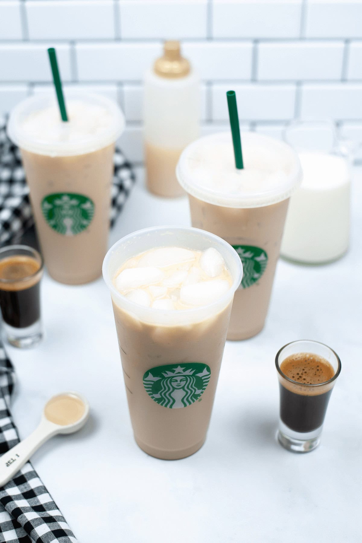 Three iced coffee drinks in Starbucks cups are placed on a white surface, including the decadent Iced White Chocolate Mocha, surrounded by two small espresso shots, a bottle of syrup, a glass of milk, a measuring spoon, and a black and white checkered cloth.
