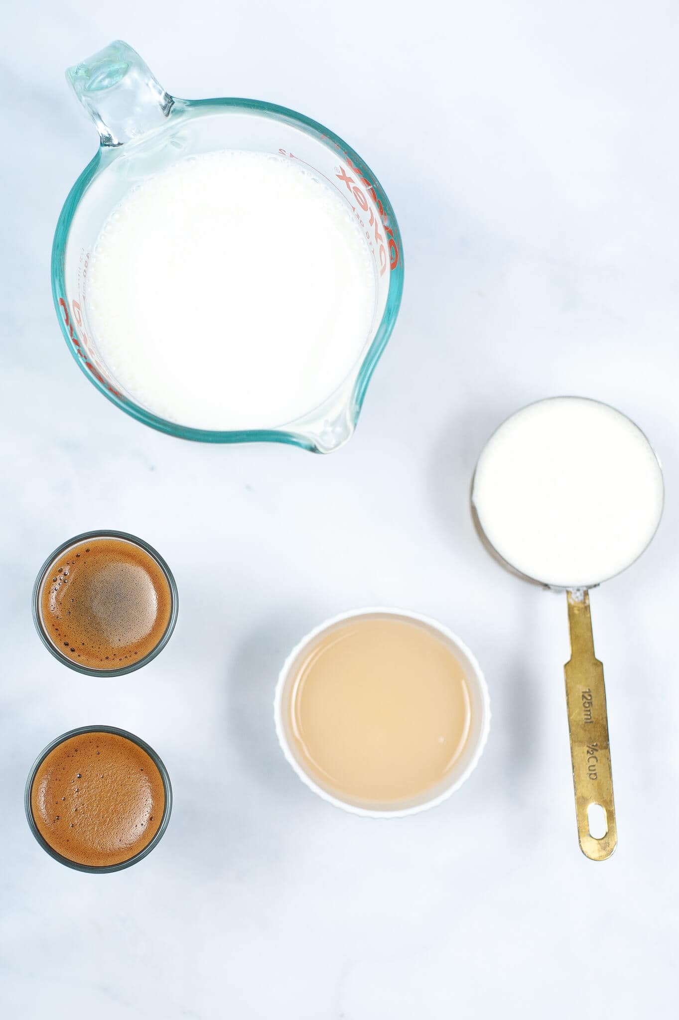 Top-down view of coffee, milk, and a drink mixture in various containers—including two small cups, a measuring cup, a small bowl, and a measuring spoon—laid out on a white surface. Get ready to create your own Iced White Chocolate Mocha masterpiece with these ingredients.