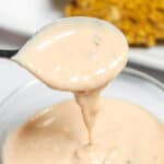 A spoon drips creamy, light orange In n Out sauce recipe into a bowl, with a blurred view of crispy fried food in the background.