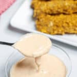 A spoonful of creamy dipping sauce, reminiscent of an In-N-Out Sauce recipe, hovers above a container, with crispy fried chicken strips arranged on a white plate in the background.