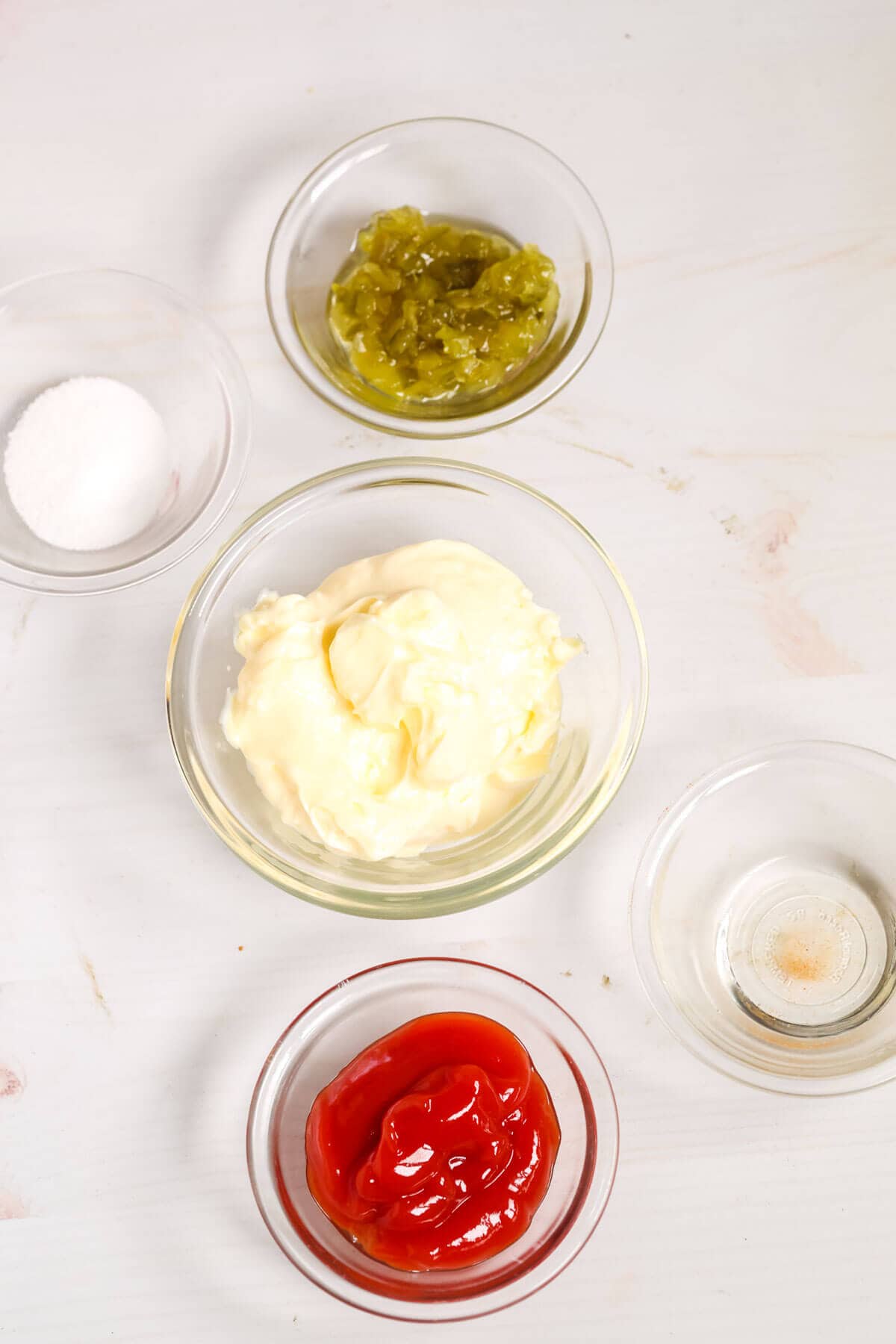 Five small glass bowls containing salt, relish, mayonnaise, ketchup, and vinegar are arranged on a light surface—key ingredients.