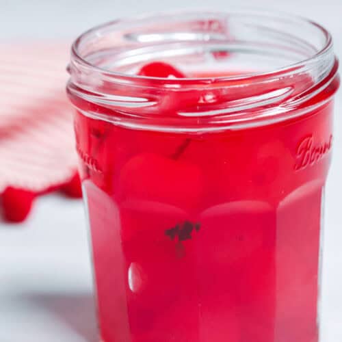 A glass jar brimming with moonshine cherries, immersed in a vibrant red liquid, sits against a light background.