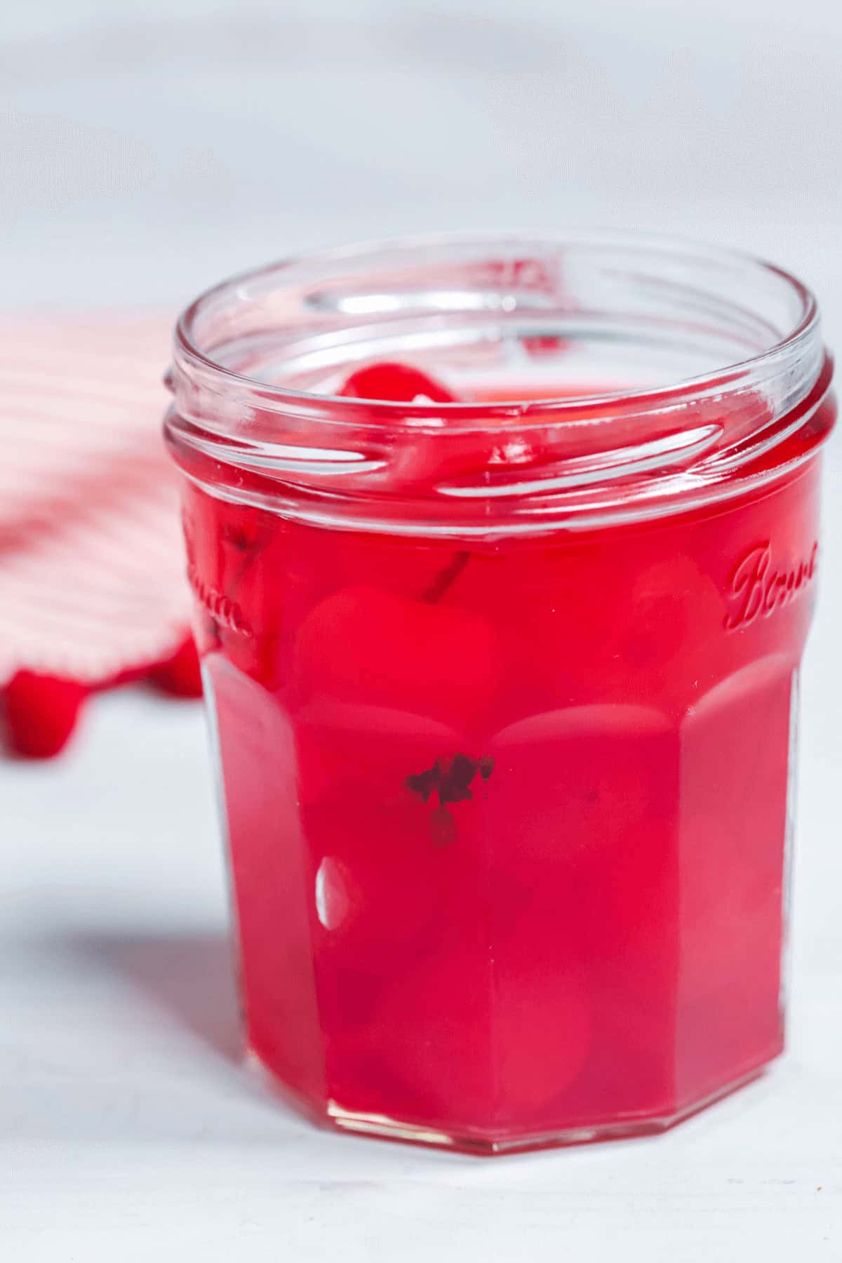 A glass jar brimming with moonshine cherries, immersed in a vibrant red liquid, sits against a light background.