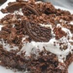 A slice of decadent Oreo Dump Cake adorned with crushed chocolate cookies and a dollop of whipped cream sits invitingly on a pristine white plate.