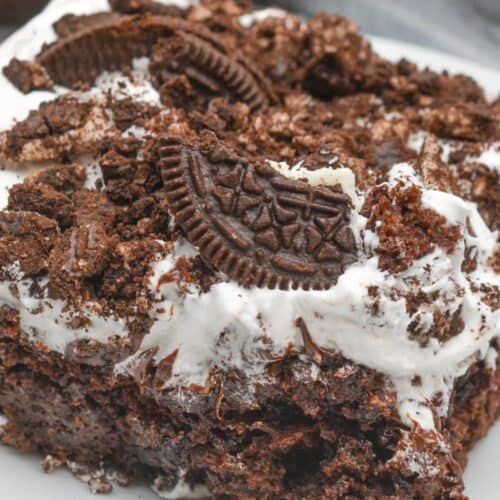 A slice of decadent Oreo Dump Cake adorned with crushed chocolate cookies and a dollop of whipped cream sits invitingly on a pristine white plate.