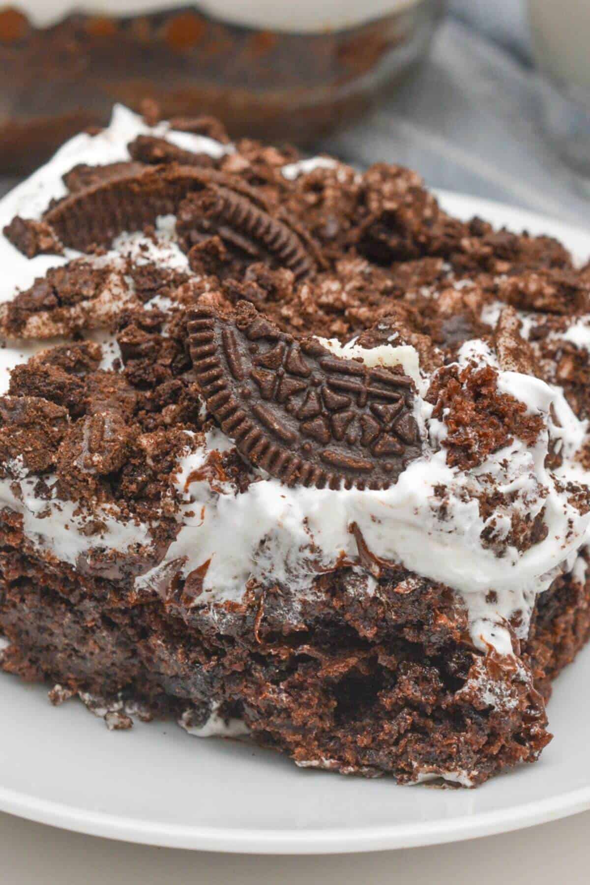 A slice of decadent Oreo Dump Cake adorned with crushed chocolate cookies and a dollop of whipped cream sits invitingly on a pristine white plate.
