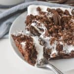 A slice of dessert adorned with crushed cookie topping and white icing sits invitingly on a plate, accompanied by a fork. In the background, cookies await alongside a glass of milk.