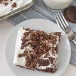 A slice of Oreo Dump Cake sits on a plate, adorned with crumbled cookies and white frosting. Nearby, a fork, an Oreo cookie, and a glass of milk rest on a light gray cloth.