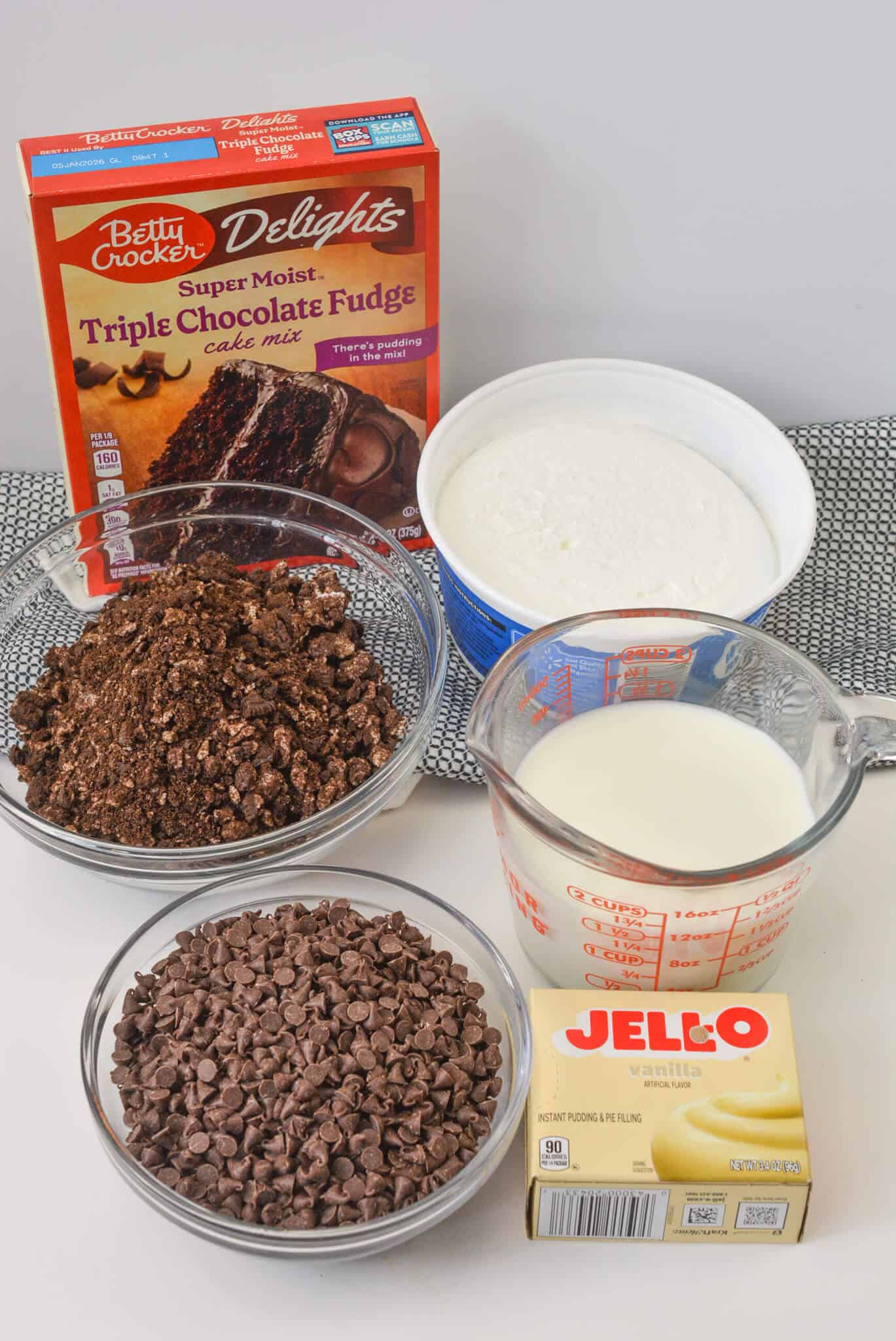 A box of triple chocolate fudge cake mix sits beside a bowl of crumbled cake and chocolate chips, a carton of whipped topping, a measuring cup of milk, and a box of vanilla pudding mix—all essential ingredients for crafting the ultimate Oreo Dump Cake delight.
