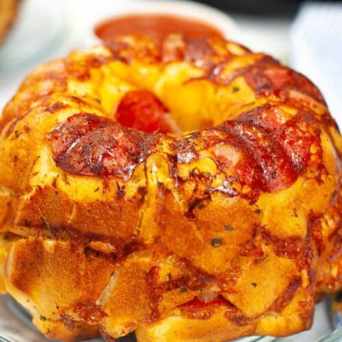 A Pizza Pull Apart Bread ring, baked to perfection with pepperoni and melted cheese, sits on a clear glass plate.