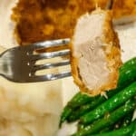 A fork holds a piece of juicy, meat against a plate with creamy mashed potatoes, crisp green beans, and another golden-brown serving of the delectable dish.