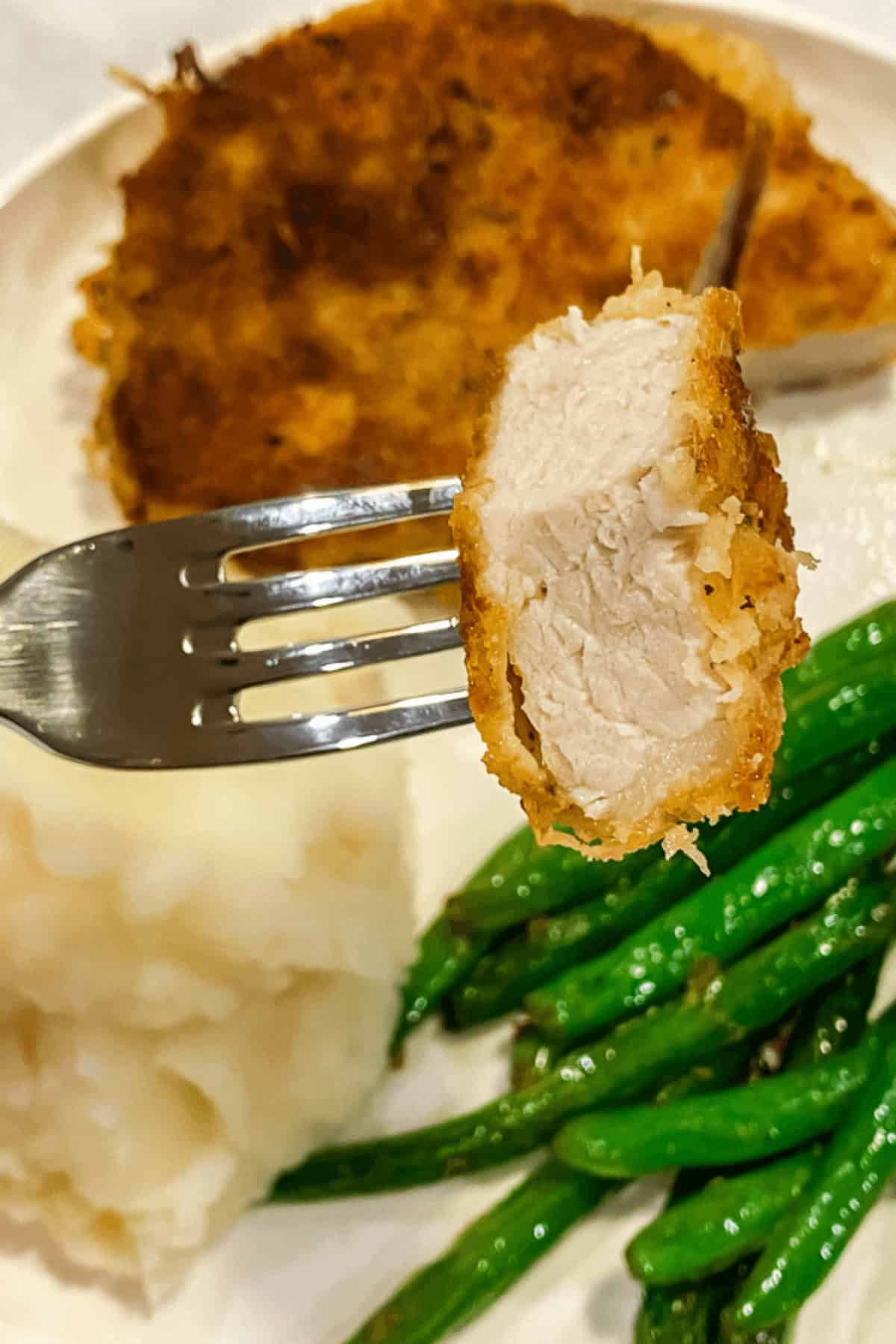 A fork holds a piece of juicy, meat against a plate with creamy mashed potatoes, crisp green beans, and another golden-brown serving of the delectable dish.