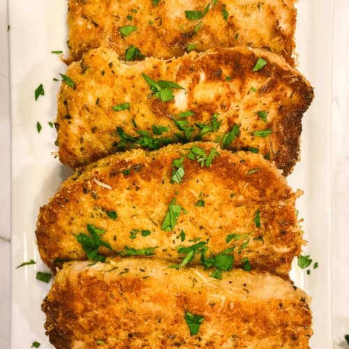 Four golden-brown breaded chicken cutlets, reminiscent of delightful baked breaded pork chops, are garnished with chopped parsley on a white rectangular plate.