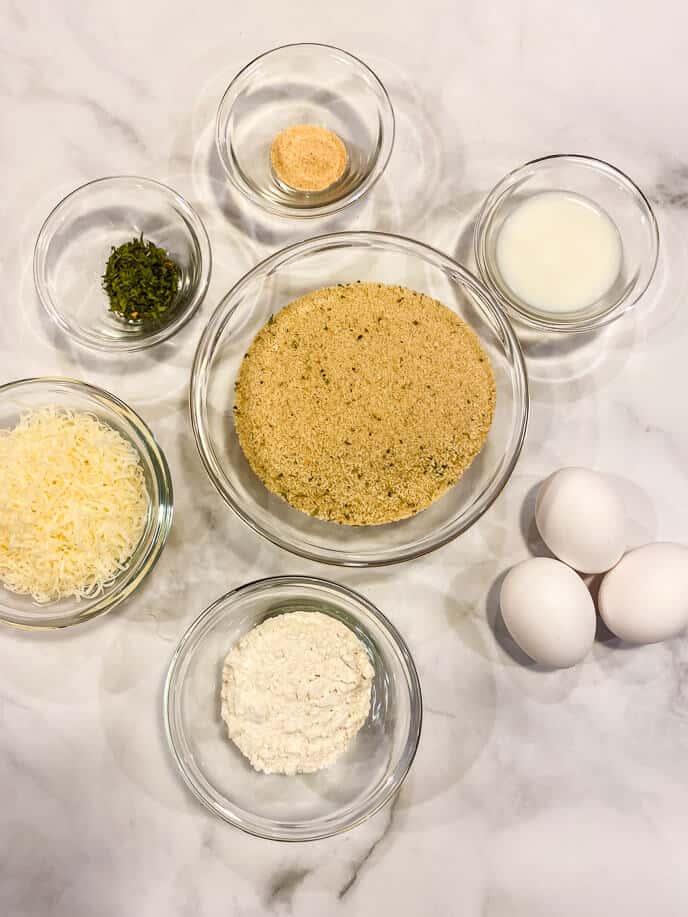 Seven bowls on a marble surface hold the perfect ingredients: breadcrumbs, grated cheese, flour, eggs, milk, spices, and garlic powder.