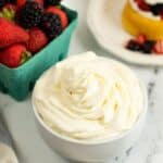 A bowl of Cream Cheese Whipped Cream sits on a marble surface next to a container of strawberries and blackberries and a dessert topped with fruit.