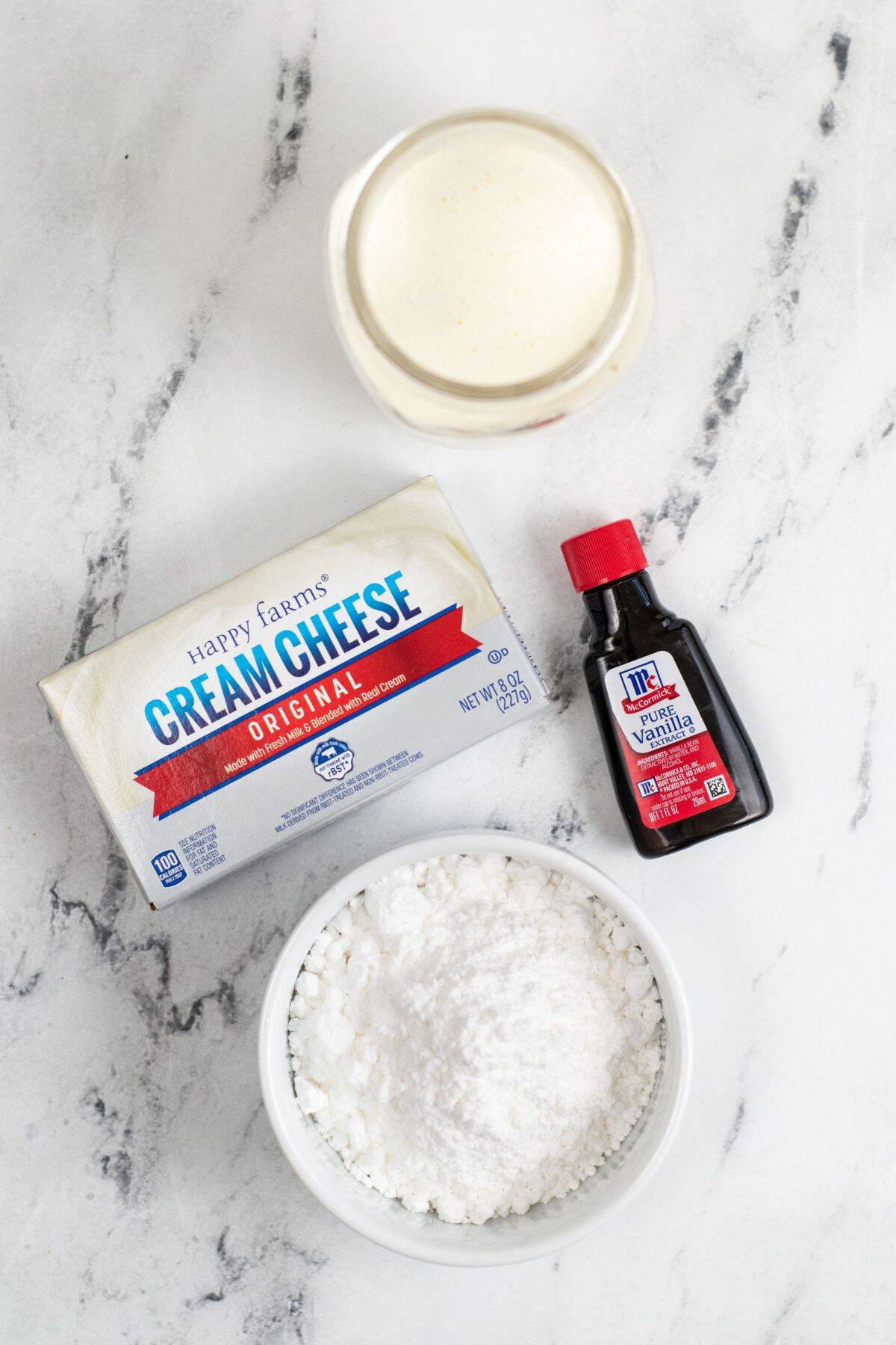A glass of milk, a block of cream cheese, a bottle of vanilla extract, and a bowl of powdered sugar on a marble surface—everything you need to make luscious Cream Cheese Whipped Cream.