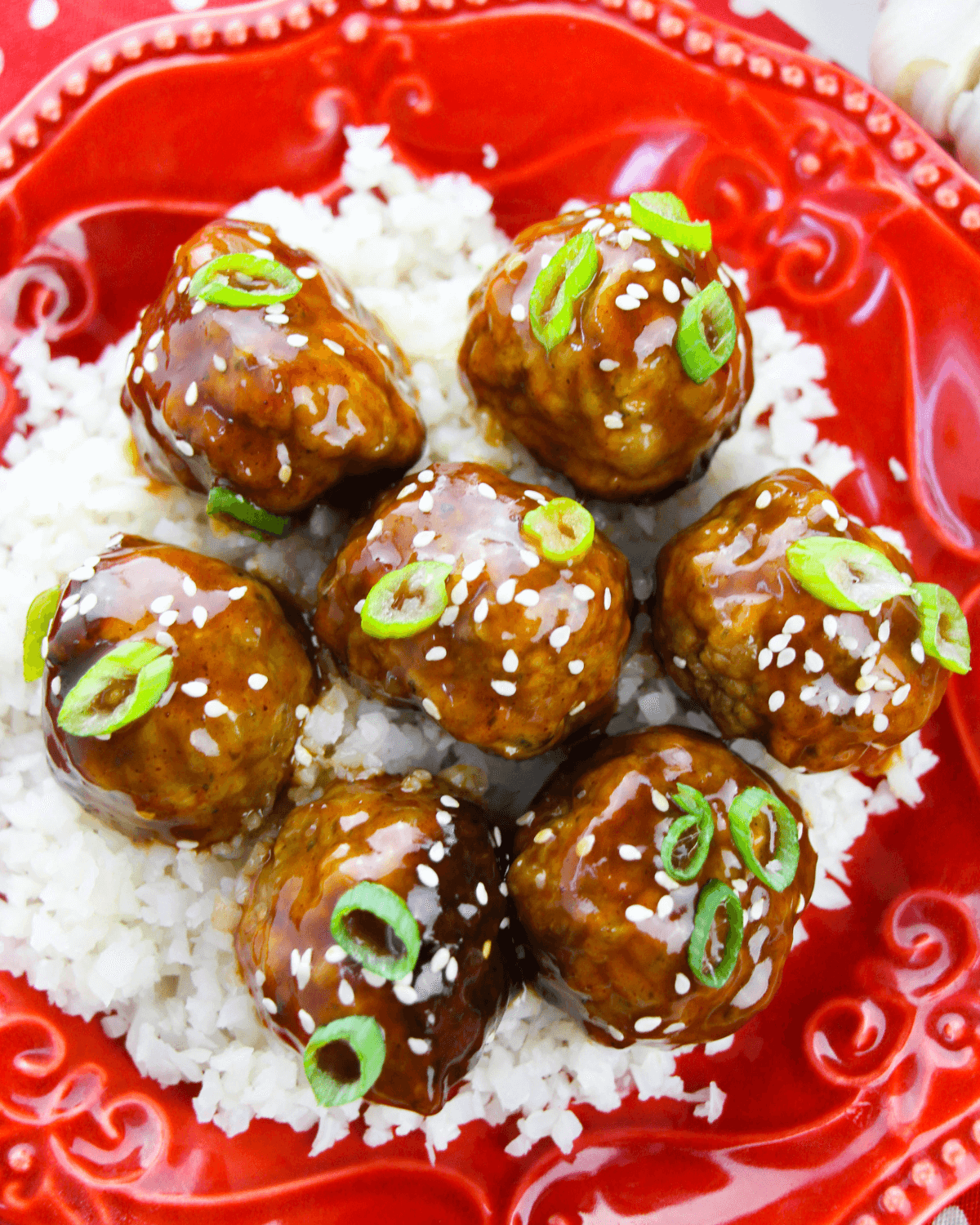 Glazed Honey Garlic Meatballs over rice garnished with sesame seeds and sliced green onions on a red plate.