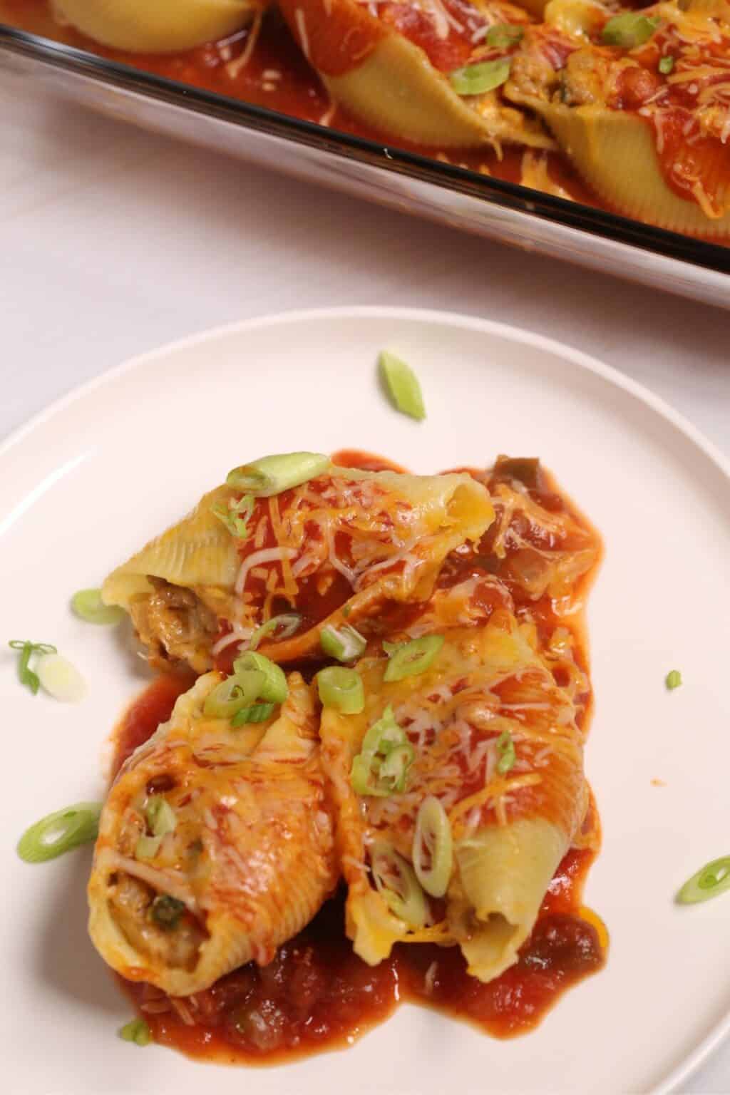 Three Mexican Stuffed Shells topped with tomato sauce, shredded cheese, and sliced green onions on a white plate, with more shells in a baking dish in the background.