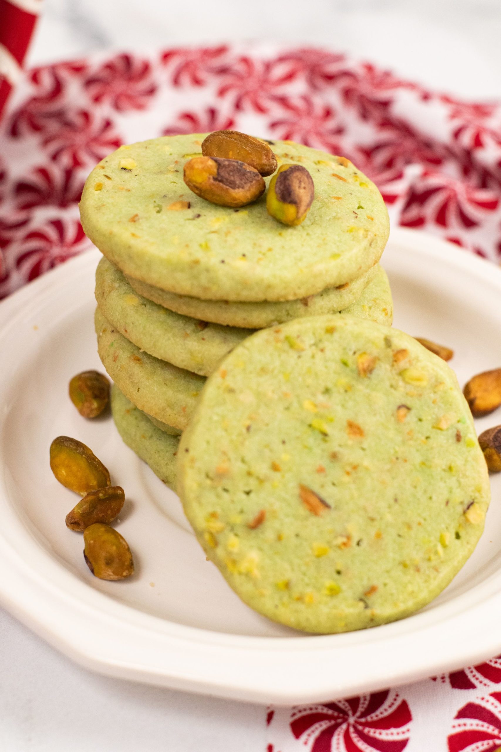 A stack of pistachio cookies on a white plate with shelled pistachios on the side