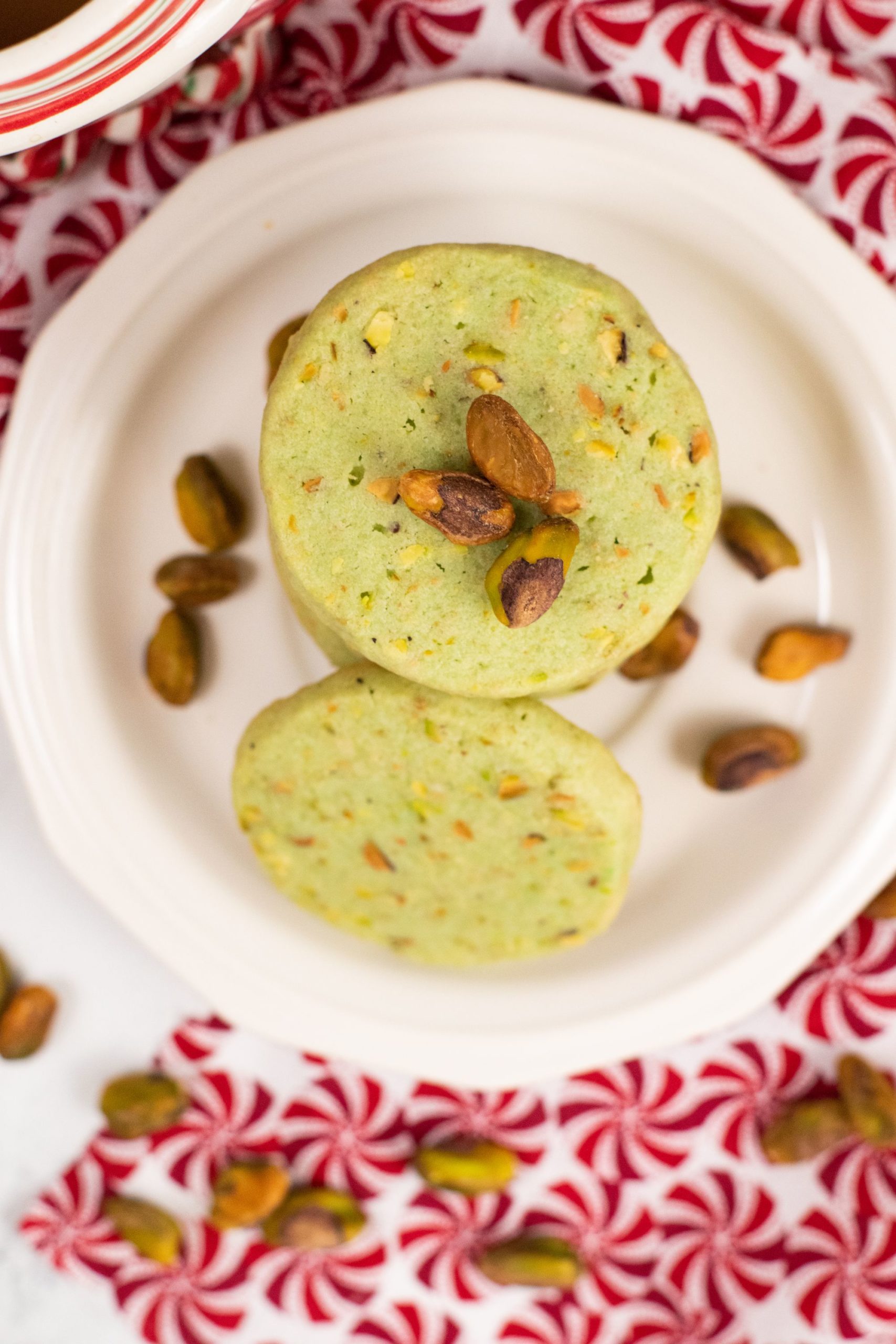 A stack of pistachio cookies on a white plate with shelled pistachios on the side