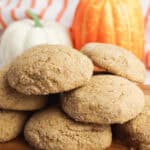 A stack of brown pumpkin spice cookies rests on a wooden board, accompanied by an orange and a white pumpkin in the background.