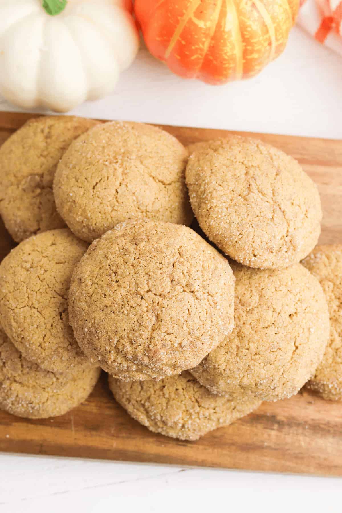 A stack of pumpkin spice cookies on a wooden board with white and orange pumpkins in the background.