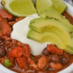 A hearty bowl of three bean chili with beans and tomatoes, topped with avocado slices, sour cream, and lime wedges.