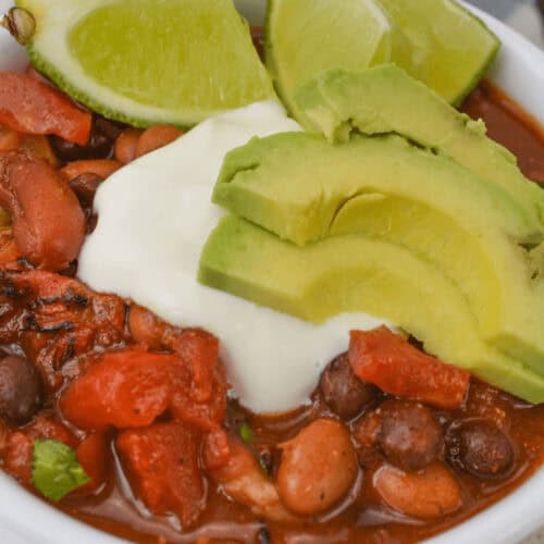 A hearty bowl of three bean chili with beans and tomatoes, topped with avocado slices, sour cream, and lime wedges.