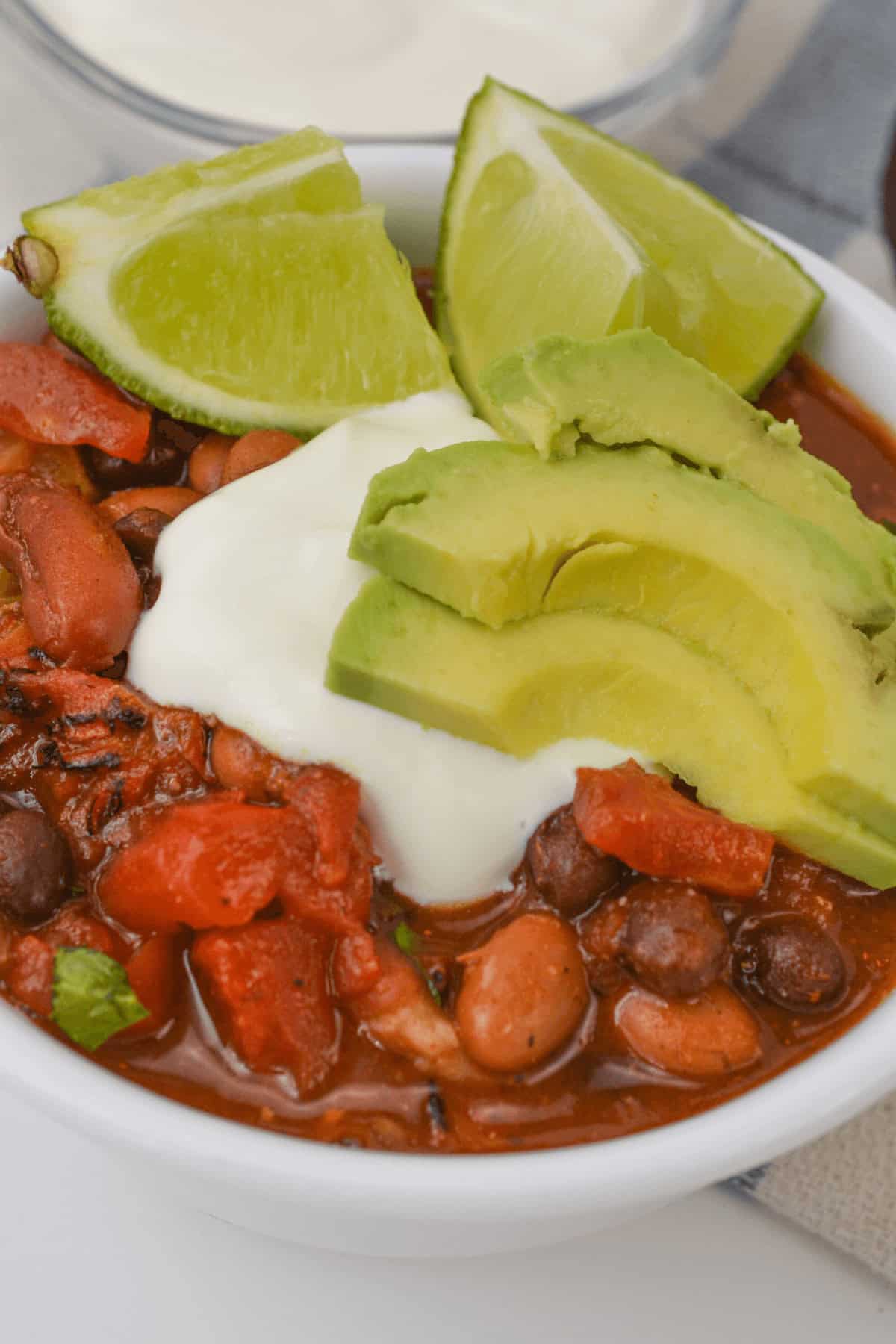 A hearty bowl of three bean chili with beans and tomatoes, topped with avocado slices, sour cream, and lime wedges.