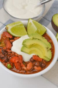 A bowl of three bean chili, topped with sliced avocado, lime wedges, and a dollop of sour cream. A bowl of sour cream and half an avocado are in the background.