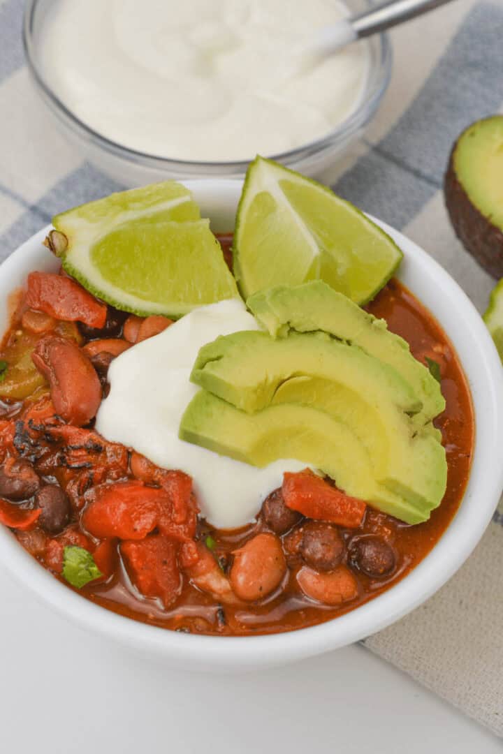 A bowl of three bean chili, topped with sliced avocado, lime wedges, and a dollop of sour cream. A bowl of sour cream and half an avocado are in the background.