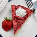 A slice of strawberry cheesecake topped with a dollop of whipped cream on a white plate next to a fresh strawberry and a fork, reminiscent of an Upside Down Strawberry Vanilla Cake.