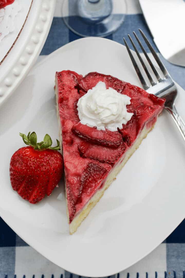 A slice of strawberry cheesecake topped with a dollop of whipped cream on a white plate next to a fresh strawberry and a fork, reminiscent of an Upside Down Strawberry Vanilla Cake.