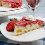 A slice of upside down strawberry vanilla cake with a dollop of whipped cream and a fresh strawberry sits on a white plate, displayed on a blue and white checkered tablecloth. A cake stand and strawberries can be seen in the background.