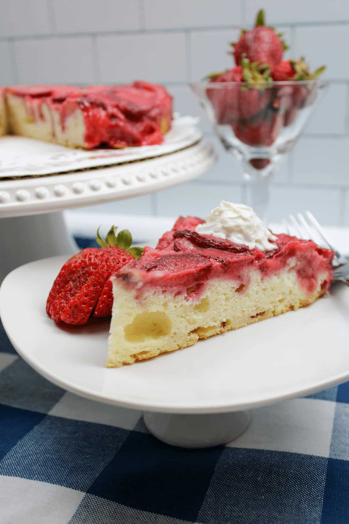 A slice of upside down strawberry vanilla cake with a dollop of whipped cream and a fresh strawberry sits on a white plate, displayed on a blue and white checkered tablecloth. A cake stand and strawberries can be seen in the background.