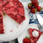 A delectable upside-down strawberry vanilla cake on a white platter with a lace doily, accompanied by a slice of the cake on a plate topped with whipped cream and a fresh strawberry, and complemented by a glass of strawberries on the side.