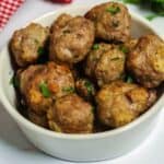 A bowl of baked meatballs garnished with chopped herbs sits on a white surface. In the background, a red and white checkered cloth and green leaves add a charming touch to this delicious meatballs recipe.