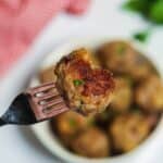 A fork holds a meatball with a bite taken out, over a plate of more baked delights. A red checkered cloth and fresh herbs in the background complete this mouthwatering scene, ideal for showcasing your favorite baked meatballs recipe.
