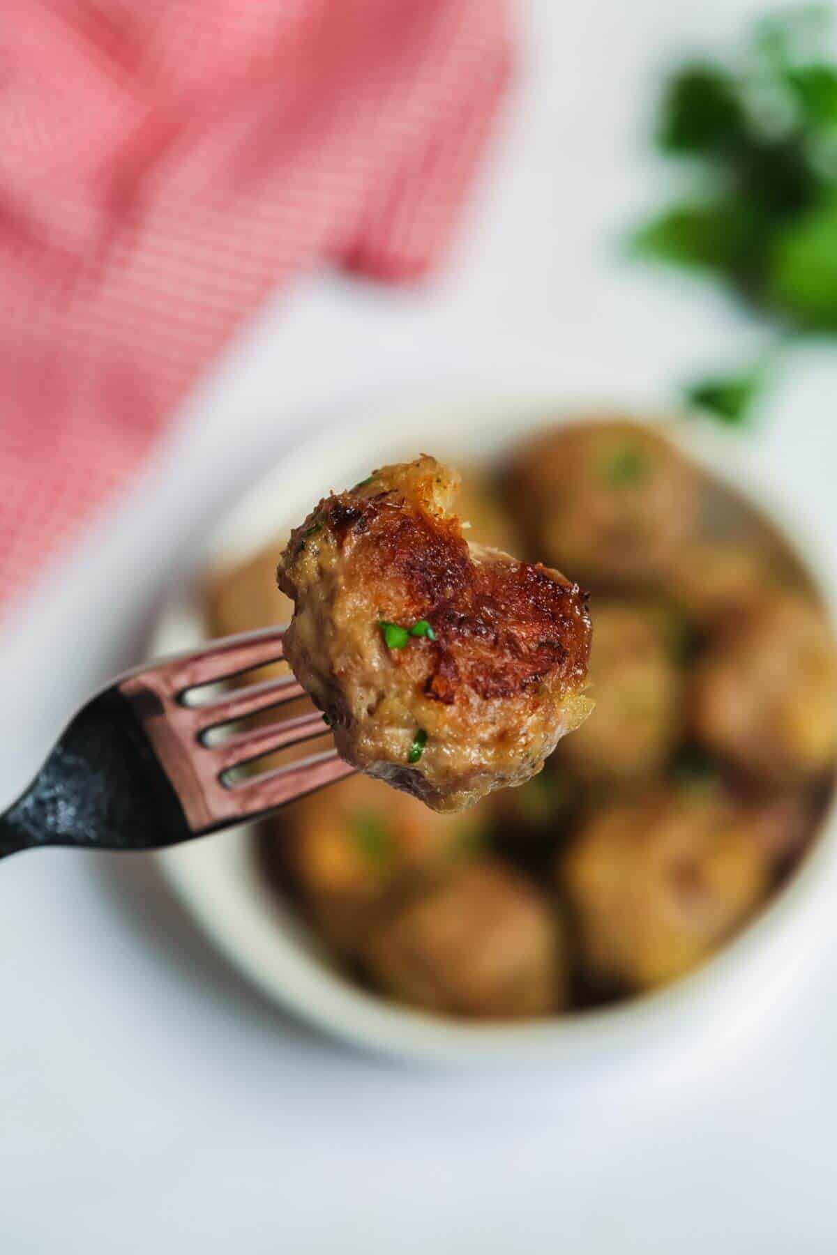 A fork holds a meatball with a bite taken out, over a plate of more baked delights. A red checkered cloth and fresh herbs in the background complete this mouthwatering scene, ideal for showcasing your favorite baked meatballs recipe.